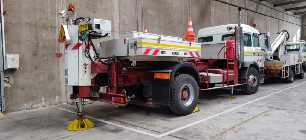 Camion de mesures spécialisé avec nacelle et stabilisateurs jaunes.